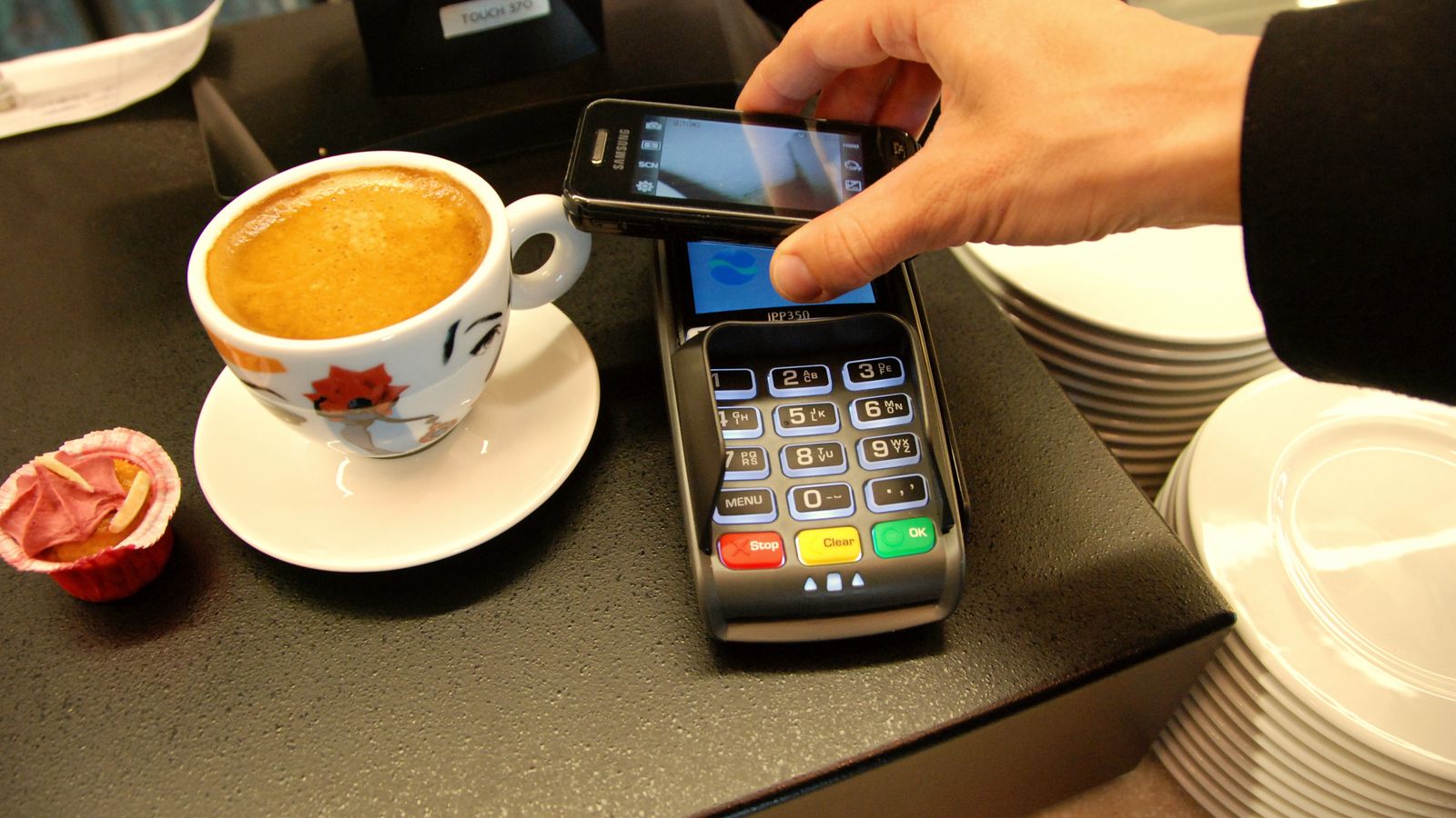 A customer holds a smartphone over a card terminal to pay at a café counter