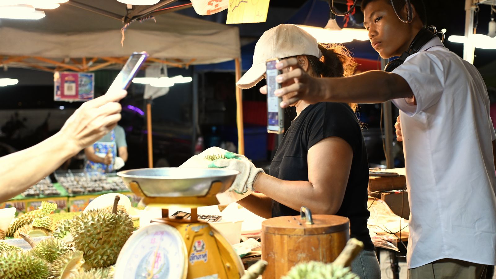 A young buyer holds up his phone to pay at a night-market fruit stall while vendors weigh durians